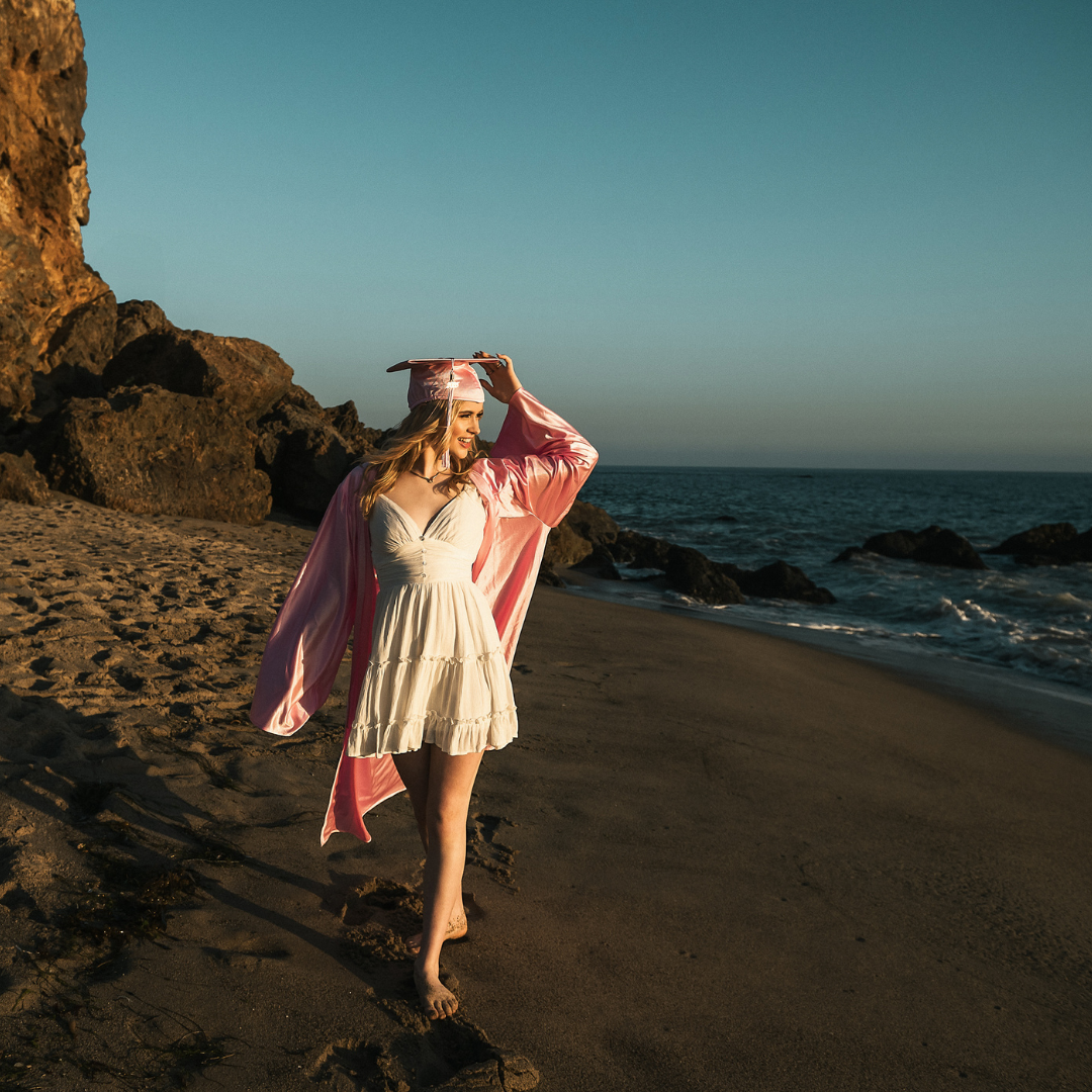 high school senior girl in a white dress and a pink cap and gown at the beach in malibu for her senior portrait session