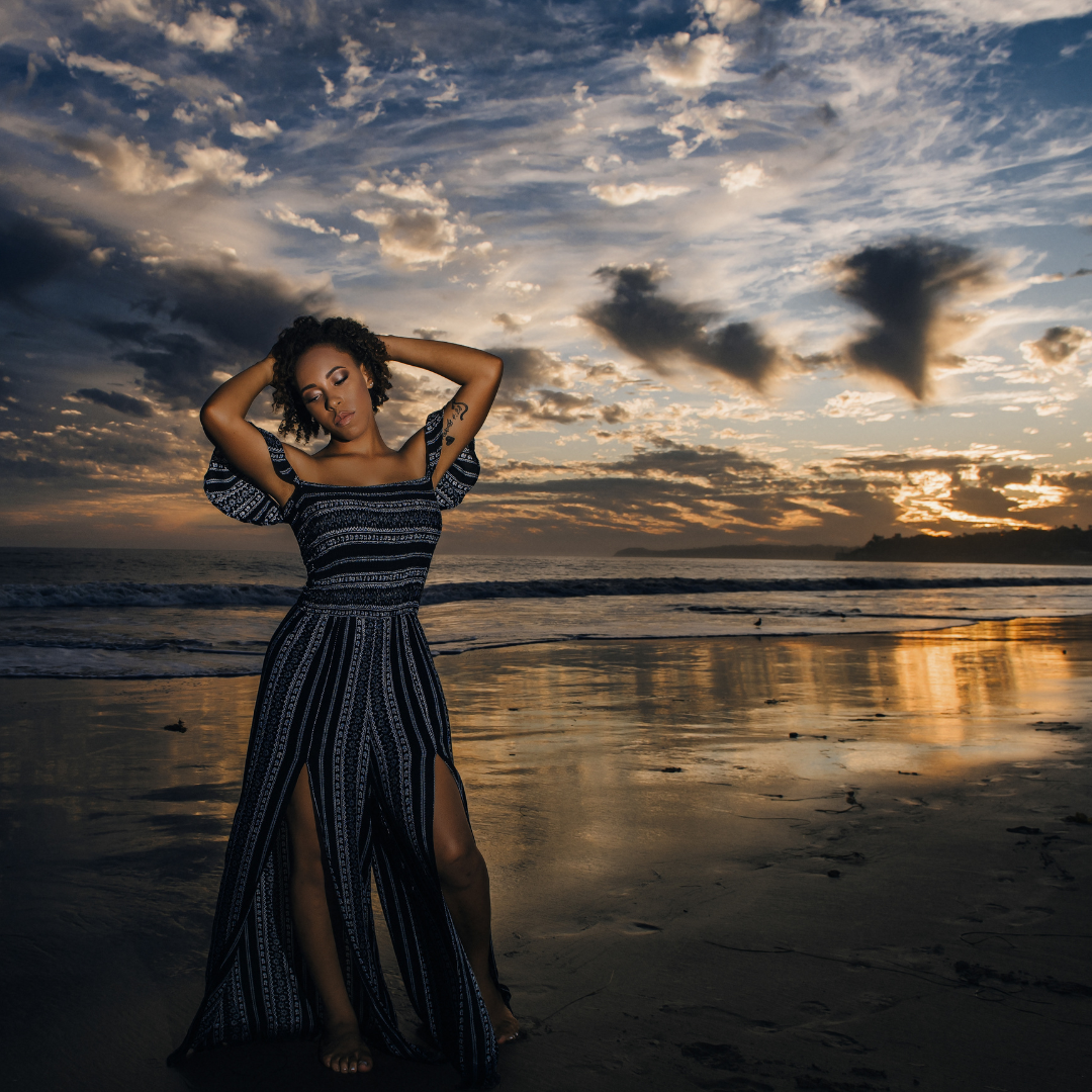 african american teen girl in a long blue dress standing on the beach with a beautiful sunset behind her, looking down the right